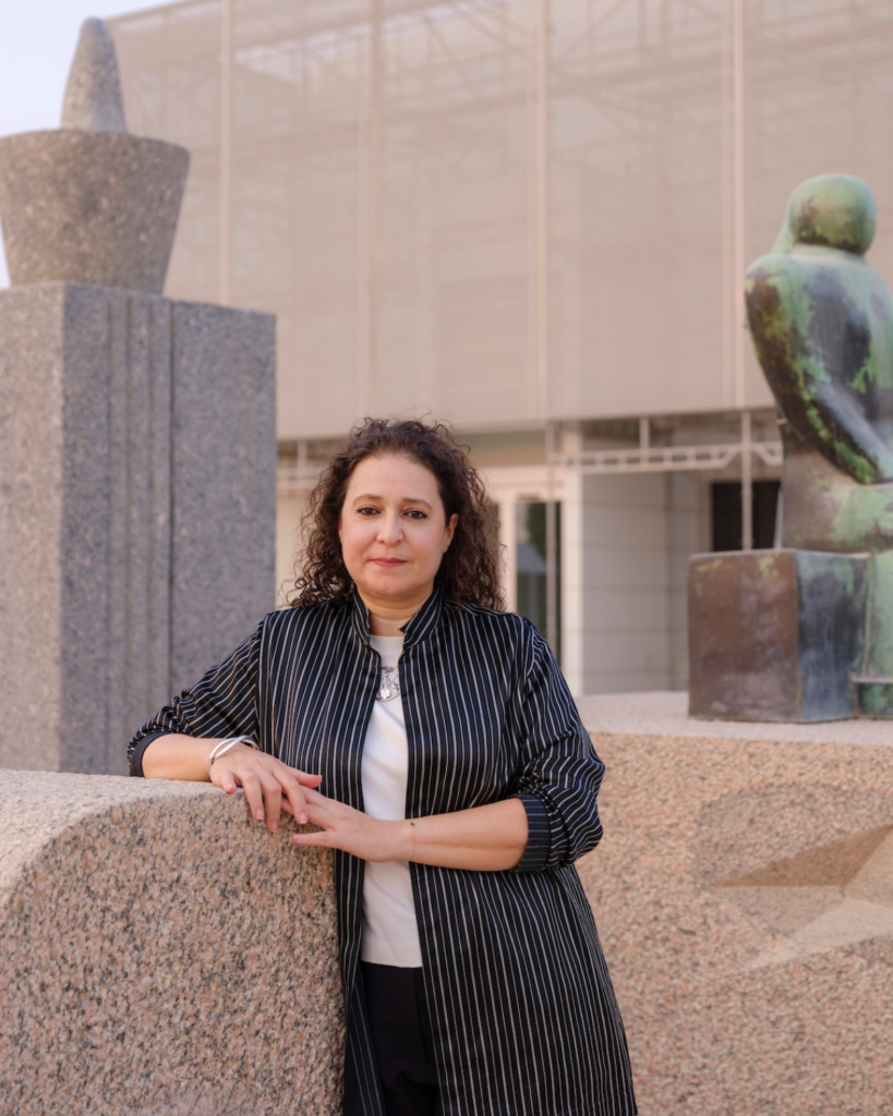 A woman with curly hair stands outdoors, leaning on a stone wall. She wears a striped jacket over a white top. Behind her are modern sculptures and a building with a glass facade.