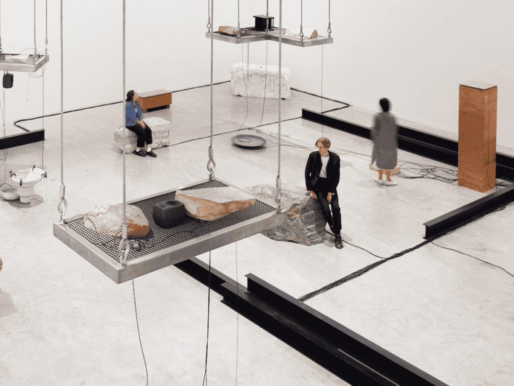Three people sit or stand among scattered rocks, platforms, and modern art installations in a spacious, minimalist gallery with white walls and a concrete floor. Suspended trays hold stones and objects overhead.