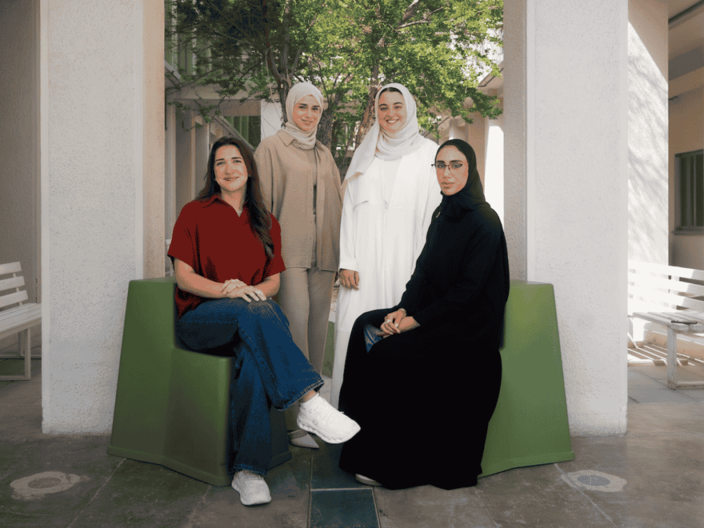 Four women pose together outdoors. Two are seated on modern green benches while two stand behind them. They wear modest clothing in neutral and dark colors, and trees and white columns frame the background.