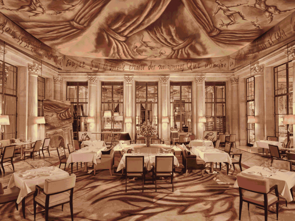 Elegant restaurant with tall columns, large windows, and tables set with white cloths; dramatic ceiling art and warm, ambient lighting create a luxurious, sophisticated atmosphere.