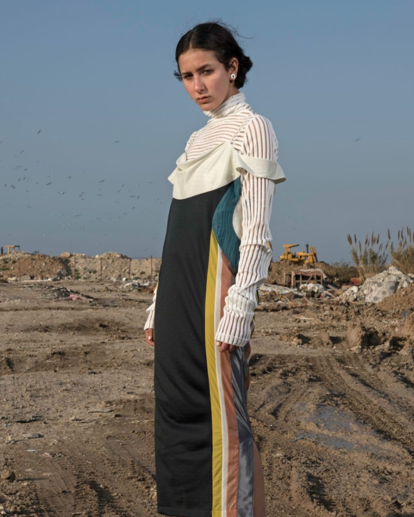 A woman in a long, color-block dress and striped, ruffled white top stands on a muddy construction site with debris, dirt, and blue sky in the background. She gazes seriously at the camera.