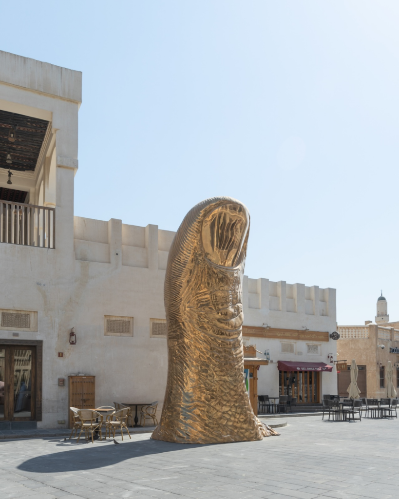 A large gold thumb sculpture stands upright in an open plaza with white stone buildings and empty outdoor tables under a clear sky.