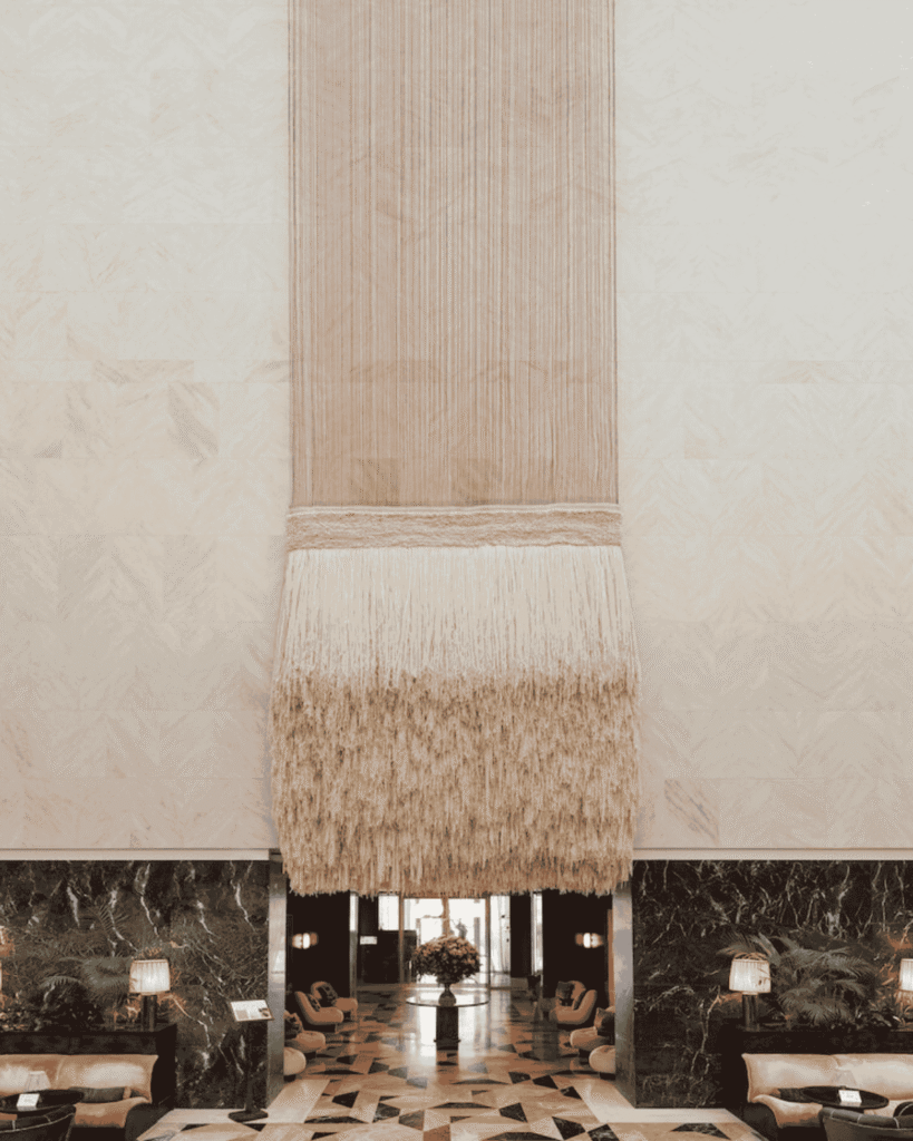A hotel lobby with a dramatic, tall beige fringe wall hanging, marble floors with geometric patterns, dark green marble walls, seating areas, and a flower arrangement near glass doors in the background.