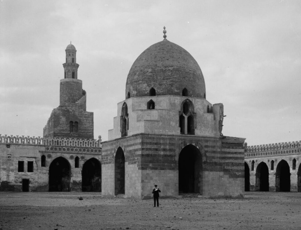 A large stone mosque with a central dome and arched entrances stands in an open courtyard. A person is visible in front of the building. A tall, spiral minaret rises in the background under a cloudy sky.
