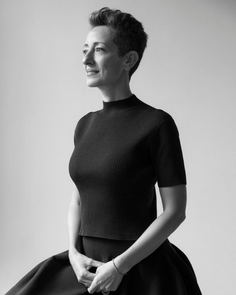 Black and white portrait of a person with short hair, wearing a ribbed, short-sleeve top and a skirt, standing and looking to the left, smiling softly with hands clasped in front.