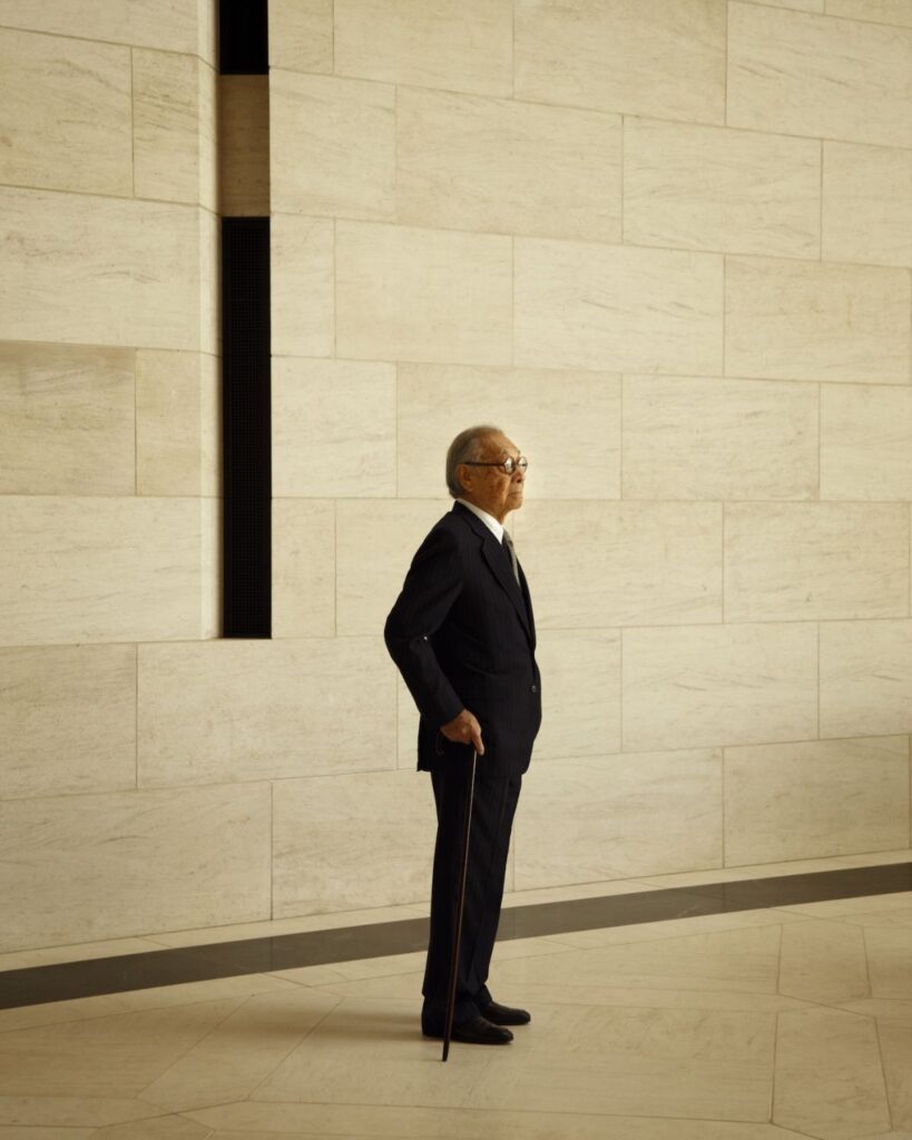 An older man in a dark suit stands in profile, holding a cane, against a large beige stone wall with geometric patterns in a modern, spacious interior.