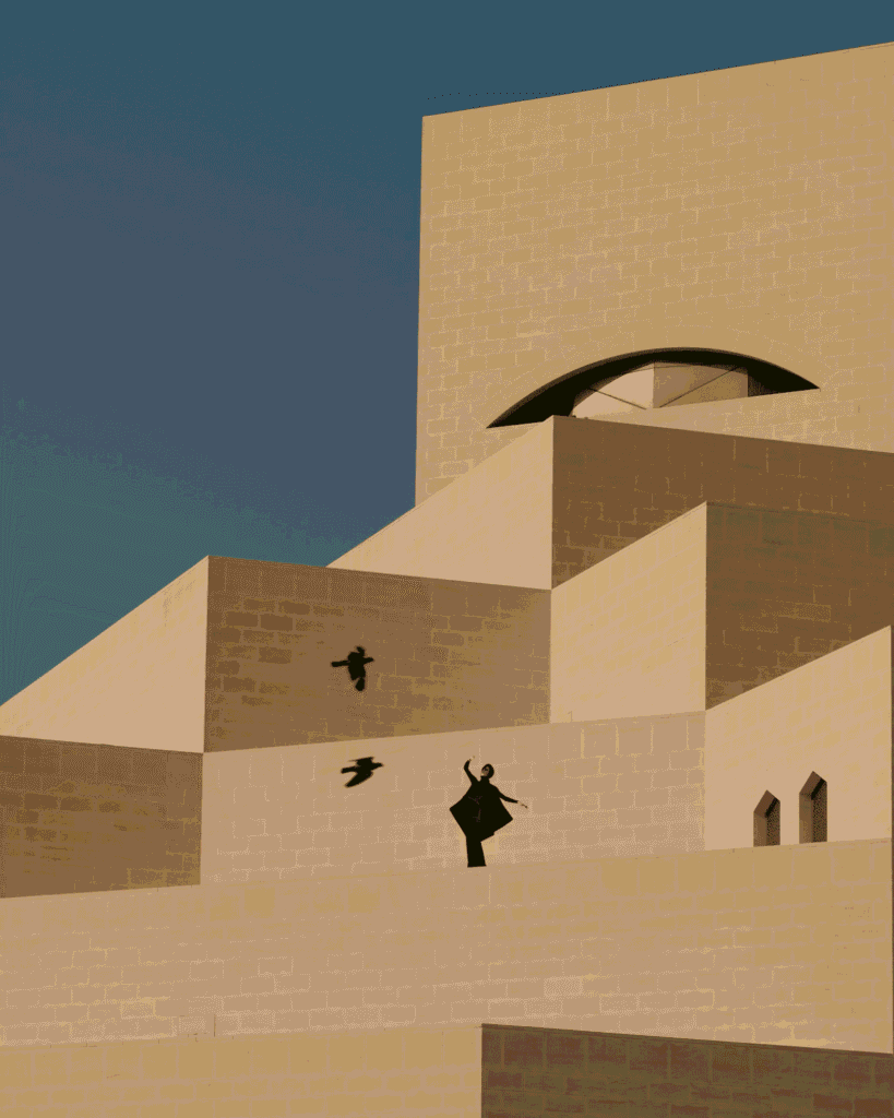 A person stands on the geometric, sand-colored steps of a modern building with an arched window, raising their arms as two birds fly nearby against a clear blue sky.