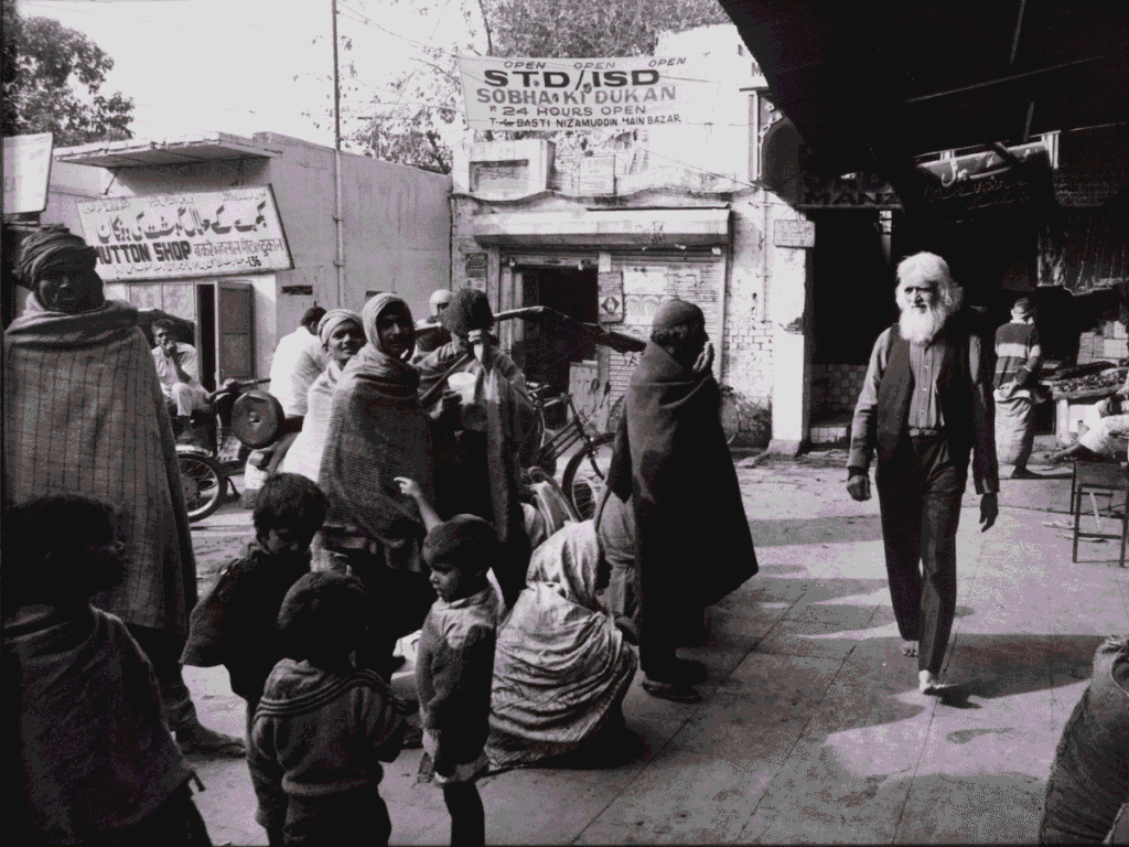 A group of people, wrapped in shawls, gathers on a busy street in front of shops. An elderly man with a long white beard walks past. Bicycles and motorcycles are visible, and signs hang above the shops.