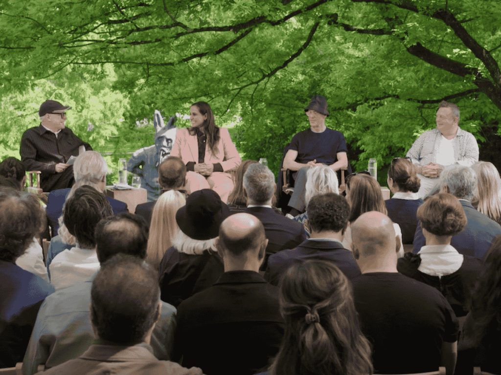 Four people sit on stage in front of a seated audience, engaged in a panel discussion outdoors beneath lush green trees. The panelists appear to be in conversation, with microphones and water glasses on the table.