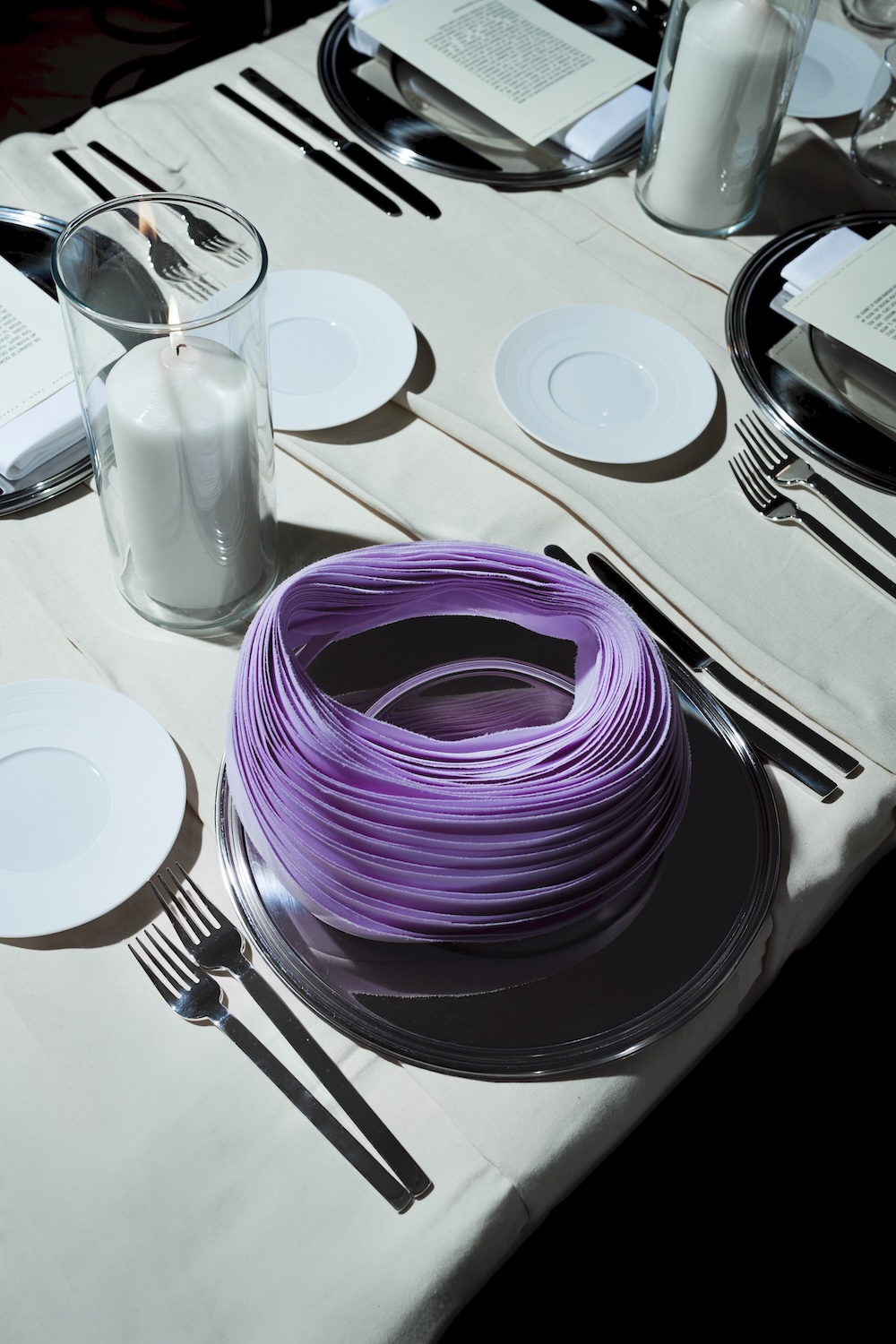 A table is set with white plates, silverware, napkins, glasses, and a large white candle. At the center, a stack of coiled purple paper sits on a plate, surrounded by formal place settings.