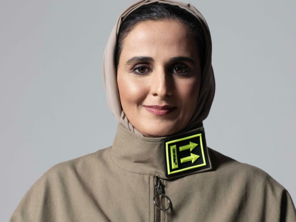 A woman wearing a beige headscarf and jacket with a yellow and black patch near the collar, featuring two arrows pointing in opposite directions, stands against a plain gray background—her style reflects the contemporary spirit of Art Basel Qatar.