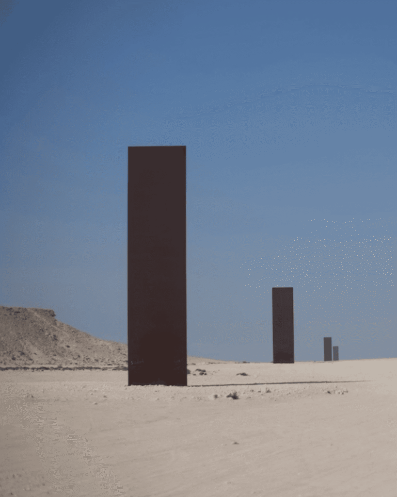 Three tall, dark monoliths stand upright in a row on a barren, sandy desert landscape under a clear blue sky. A small rocky hill appears on the left side of the image.