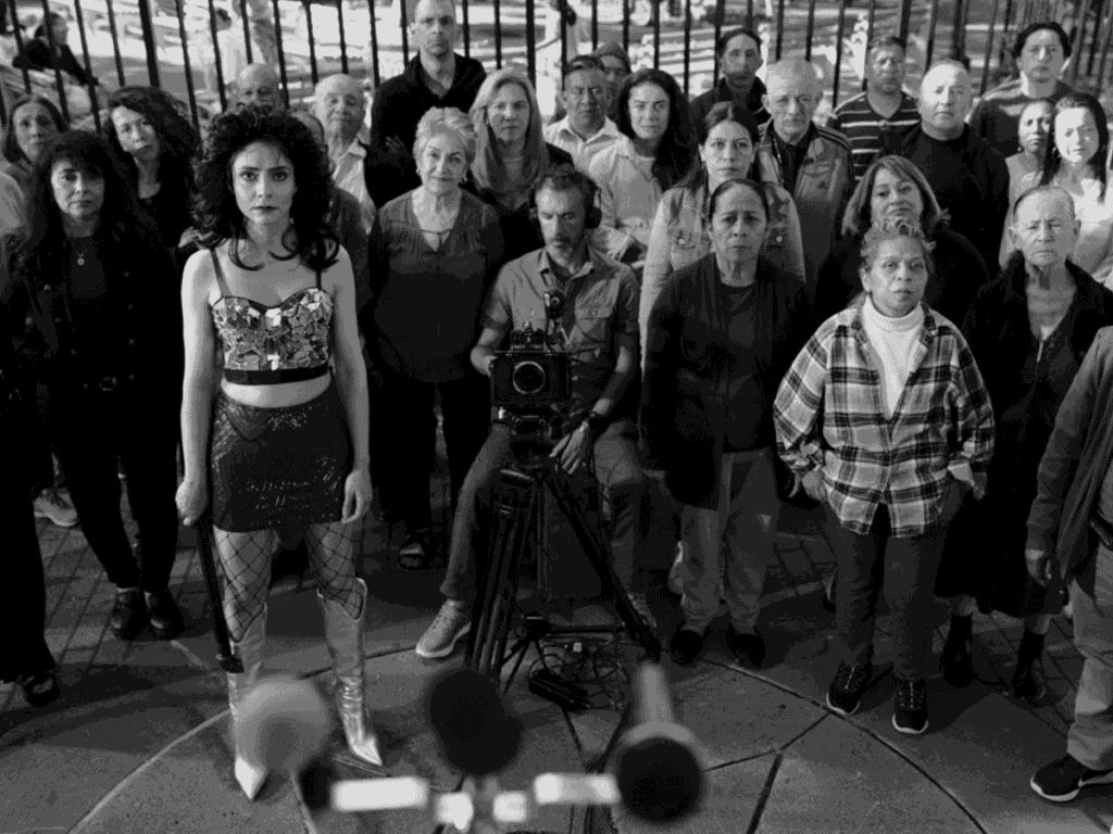A woman in a bold outfit stands in front of microphones, facing the camera, with a diverse group of serious-looking people and a person with a film camera behind her; all gaze forward in a public outdoor setting.