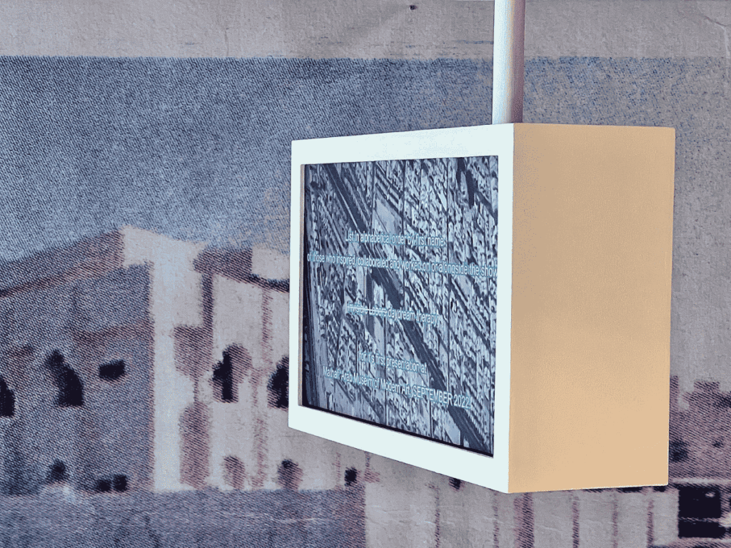 A small, white-framed digital display hangs on a wall, showing a grayscale image with architectural patterns and faint text. The background features a blurred, mural-like photo of a building with arches.