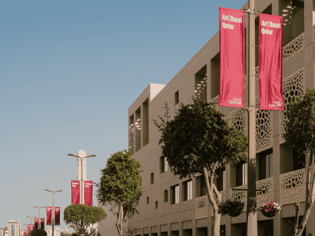 Street view of modern beige buildings with geometric patterns, lined with trees. Bright pink banners on lampposts read “Art Basel Qatar” and “Visit Qatar,” showcasing Art Basel highlights under a clear blue sky.