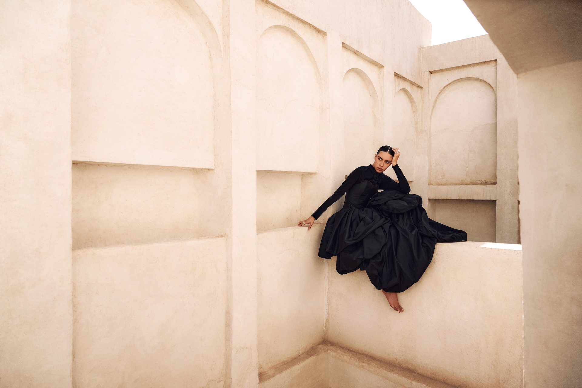 A woman in a voluminous black dress poses barefoot on a beige architectural structure with arches and soft light, evoking a dramatic scene reminiscent of Fashion Trust Arabia Prize elegance.