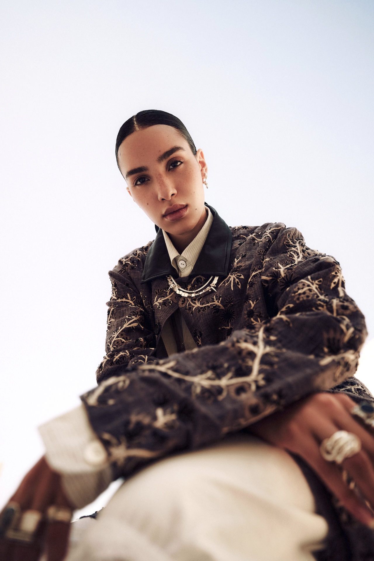 A person with slicked-back hair, wearing a detailed, embroidered jacket over a collared shirt, looks confidently at the camera from a low angle against a bright sky—a style worthy of the Fashion Trust Arabia Prize. Multiple rings adorn their fingers.