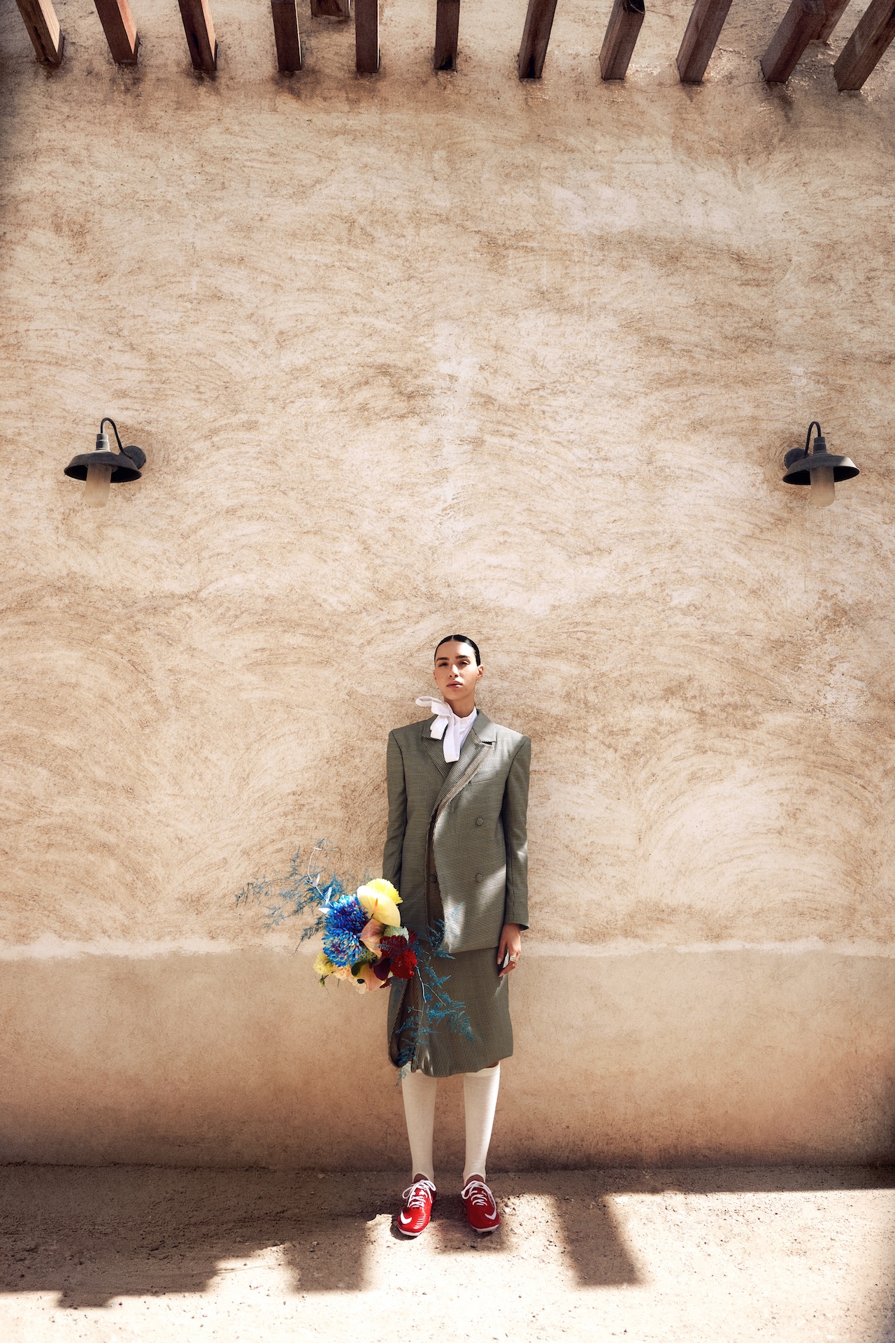 A person in a gray suit with a white blouse, white tights, and red shoes stands against a textured beige wall, holding a colorful bouquet of flowers—an image worthy of the Fashion Trust Arabia Prize. Sunlight casts shadows from beams and two black wall lamps above.