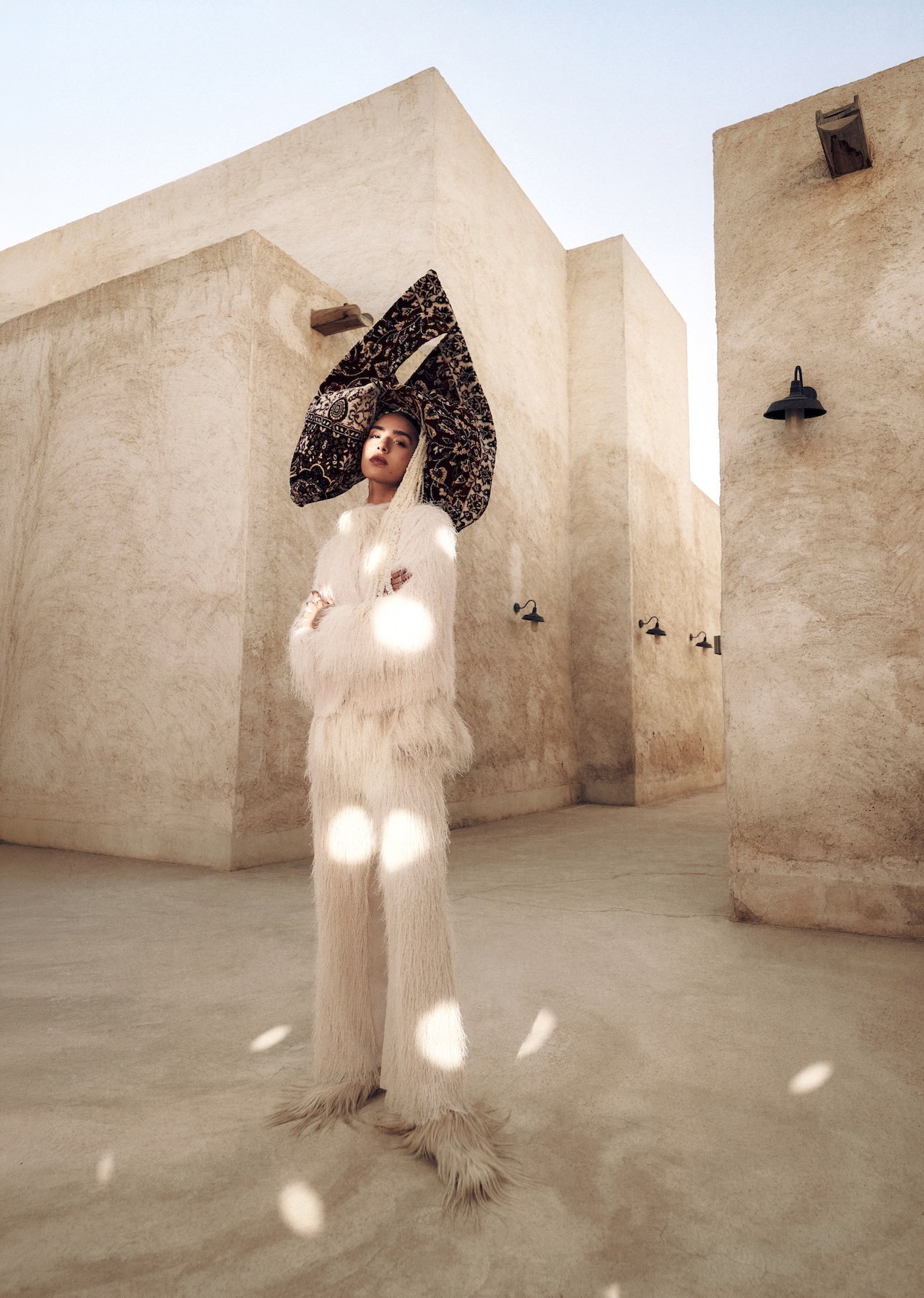 A person stands in a sunlit, beige courtyard wearing a white, furry outfit and a large, black patterned headpiece—an ensemble worthy of the Fashion Trust Arabia Prize. The geometric shadows on the walls enhance the modern and artistic atmosphere.