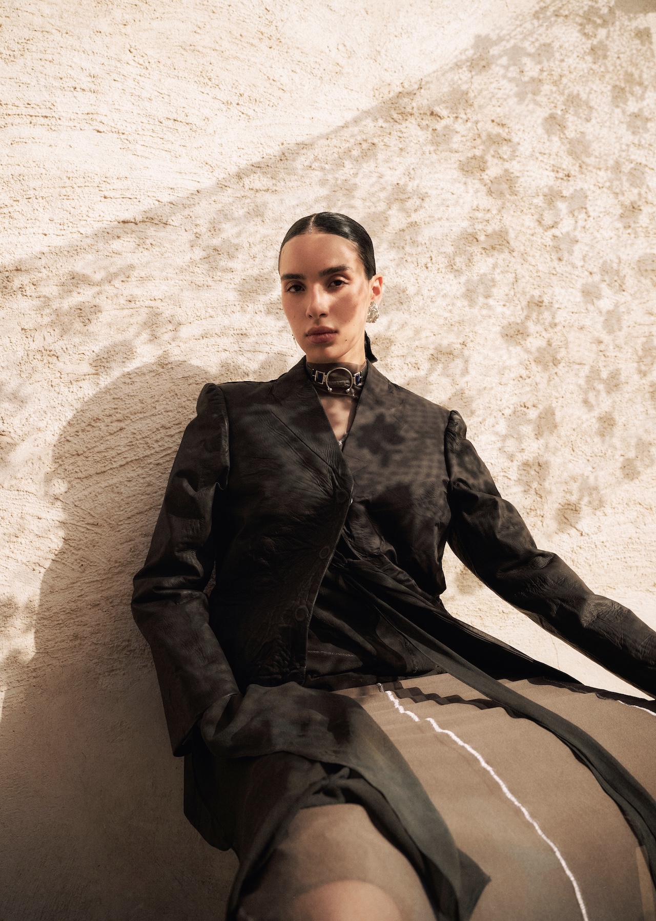 A person with dark hair tied back wears a structured black jacket and a statement choker, sitting against a textured beige wall with sunlight casting shadows—a look worthy of the Fashion Trust Arabia Prize.