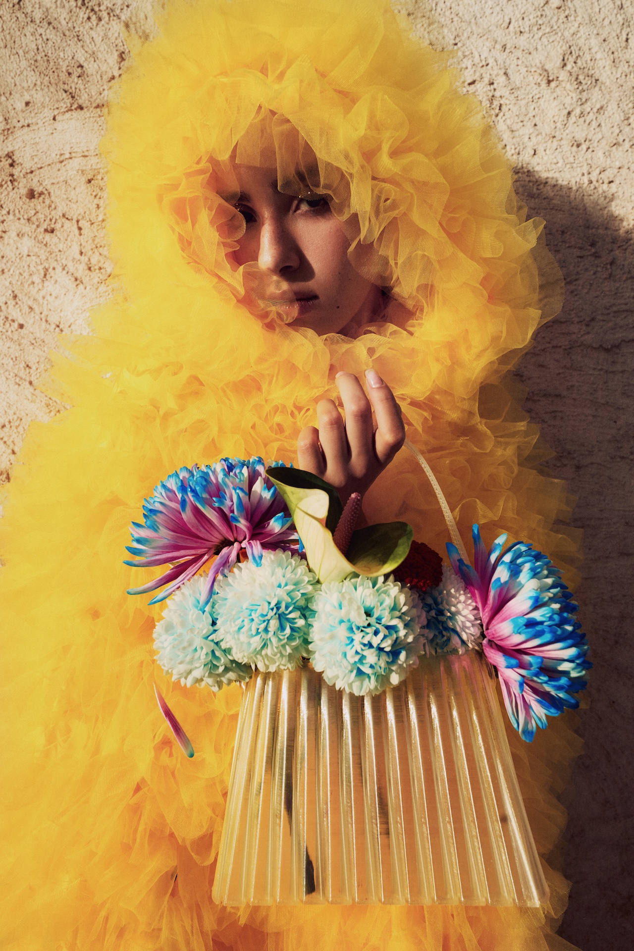A person in a bright yellow, fluffy outfit holds a clear handbag decorated with large, colorful artificial flowers, standing against a textured, light-colored wall—a look worthy of the Fashion Trust Arabia Prize.