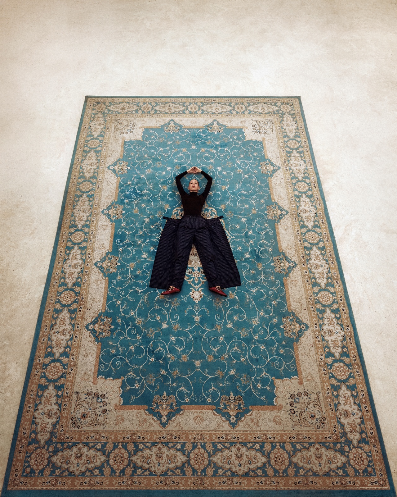 A person dressed in dark clothing, reminiscent of a Fashion Trust Arabia Prize runway look, lies on their back with arms raised above their head on an ornate blue and beige patterned rug, photographed from above on a light-colored floor.