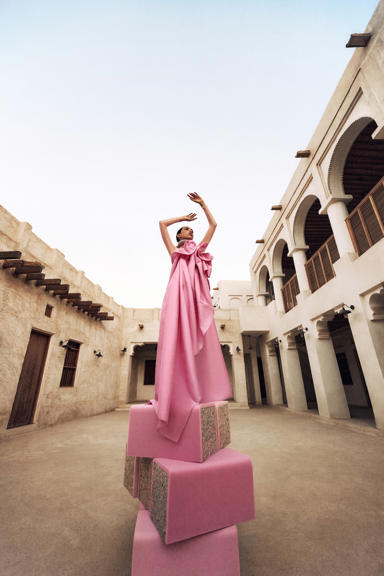 A woman wearing a long, flowing pink dress, reminiscent of the Fashion Trust Arabia Prize style, stands on stacked pink cubes in a courtyard with beige stone walls and arches, posing elegantly with her arms raised under a clear sky.
