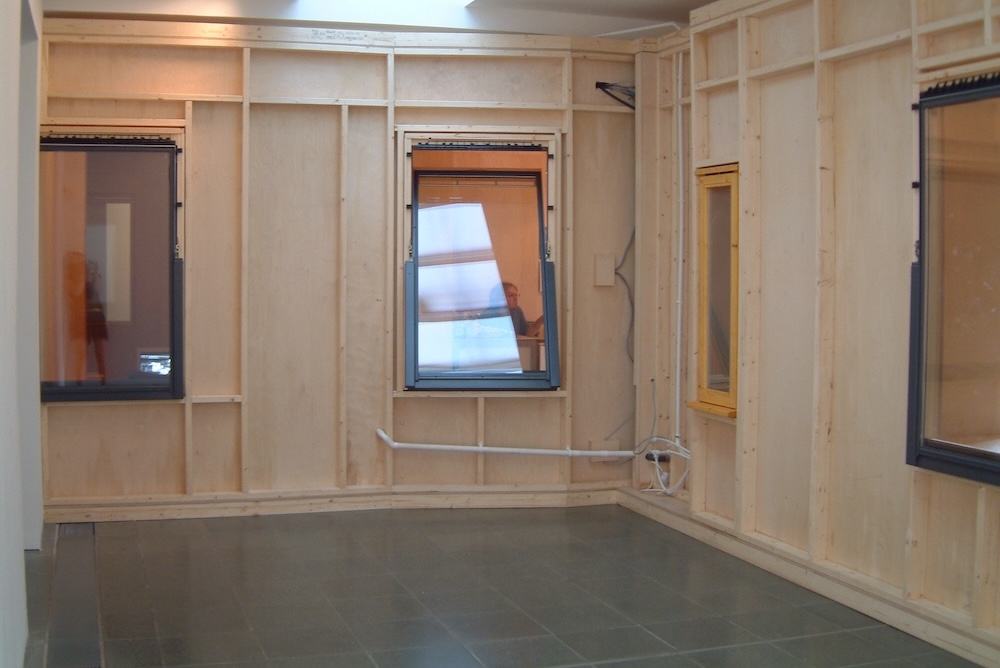 A room with unfinished wooden walls, three windows with black frames, and exposed piping along the gray tiled floor recalls a Rirkrit Tiravanija installation. Light reflects through the windows, and a person is faintly visible in one window.