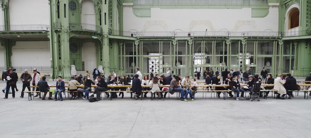 A group of people sit and socialize at long wooden tables inside a large hall with tall green metal columns and glass windows, evoking the communal spirit often found in Rirkrit Tiravanija's installations. The atmosphere appears casual and inviting.