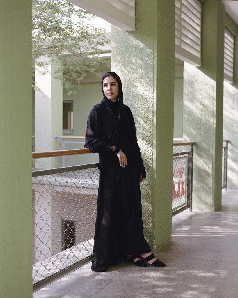 A woman wearing a black abaya and headscarf stands on a liwan balcony with light green walls and a metal railing, looking thoughtfully into the distance as natural light casts soft shadows around her.