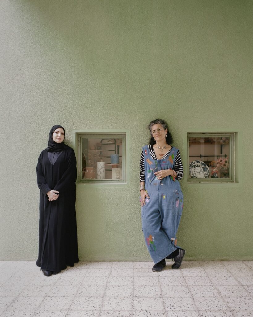 Two women stand in front of a pale green liwan wall with two small windows. One wears a black abaya and hijab, while the other wears overalls, a striped shirt, and has curly hair. Shelves with items are visible through the windows.