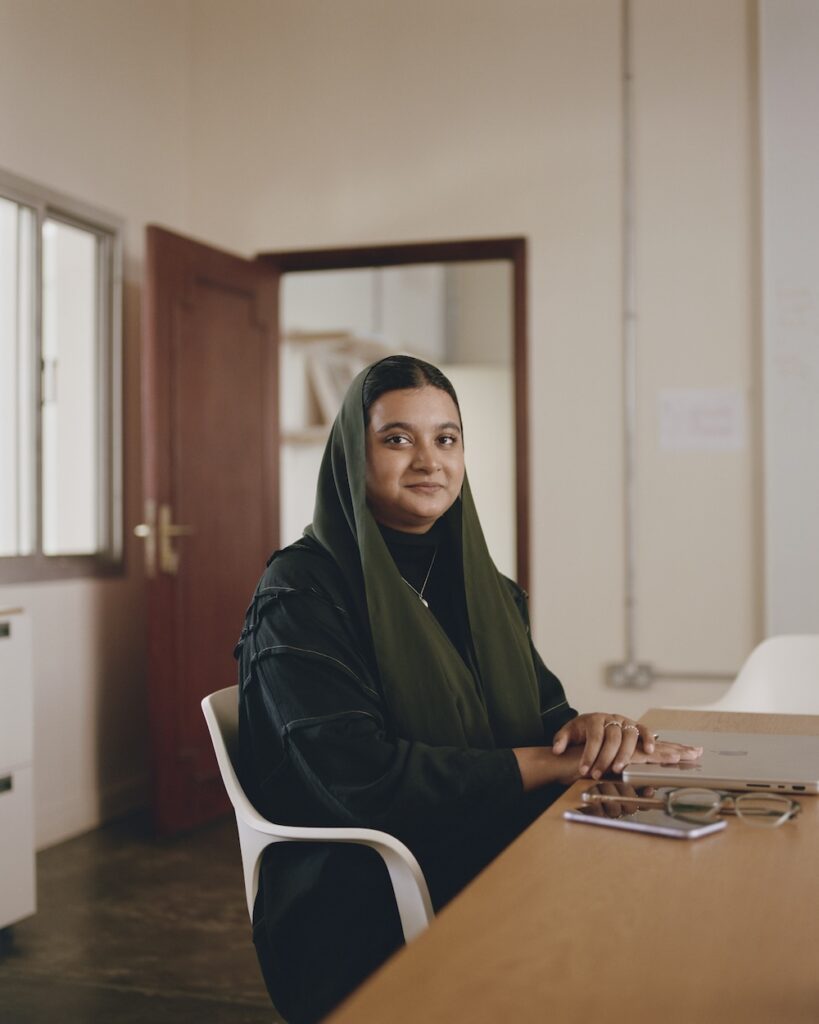 A woman wearing a dark green hijab and black clothing sits at a desk in an office liwan, looking at the camera. She has her hands folded on the desk, with a laptop and glasses in front of her. The room has a window and an open door.