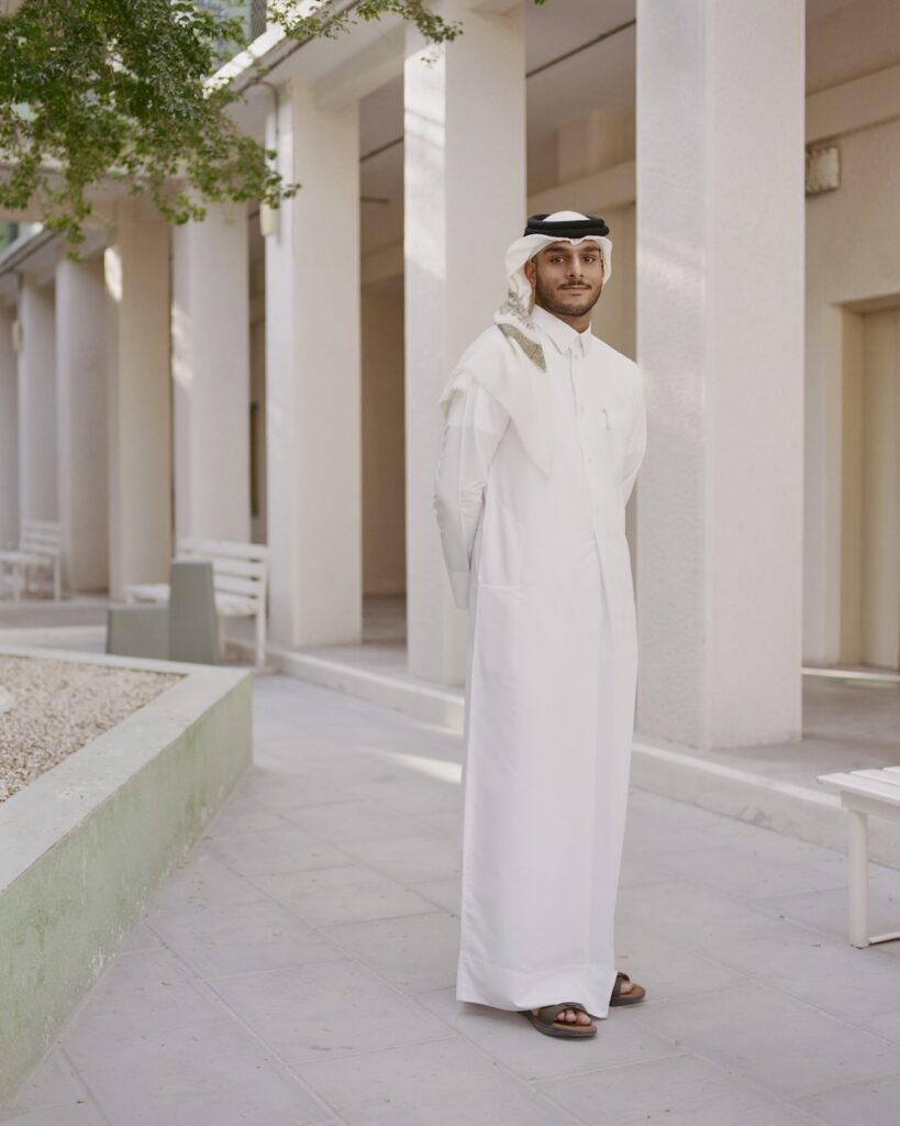 A man in traditional Middle Eastern attire stands on a shaded liwan walkway, surrounded by white columns and benches, with his hands behind his back.