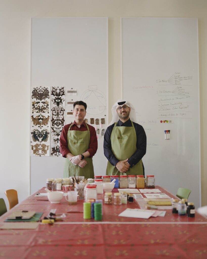 Two men wearing green aprons stand side by side in a liwan-themed classroom or art studio, behind a table with art supplies. The wall behind them features sample artwork and writing on whiteboards.