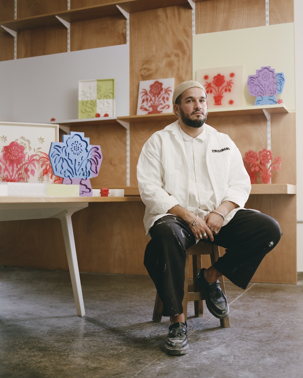 A man in a white shirt and beige beanie sits on a stool in an art studio, surrounded by colorful floral artwork inspired by liwan design, with wooden panels and a modern, creative atmosphere.