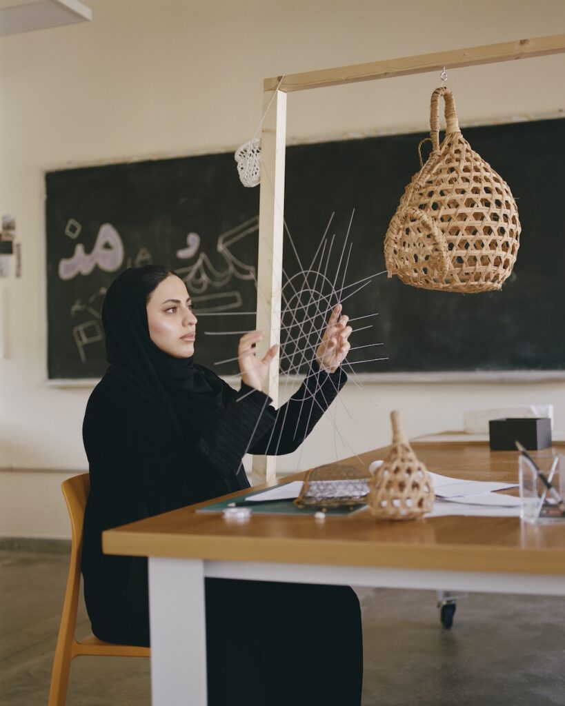 A woman in a black hijab sits at a liwan desk, weaving a net-like structure with thin threads. Woven basket lamps hang nearby, and a chalkboard with Arabic writing is in the background.