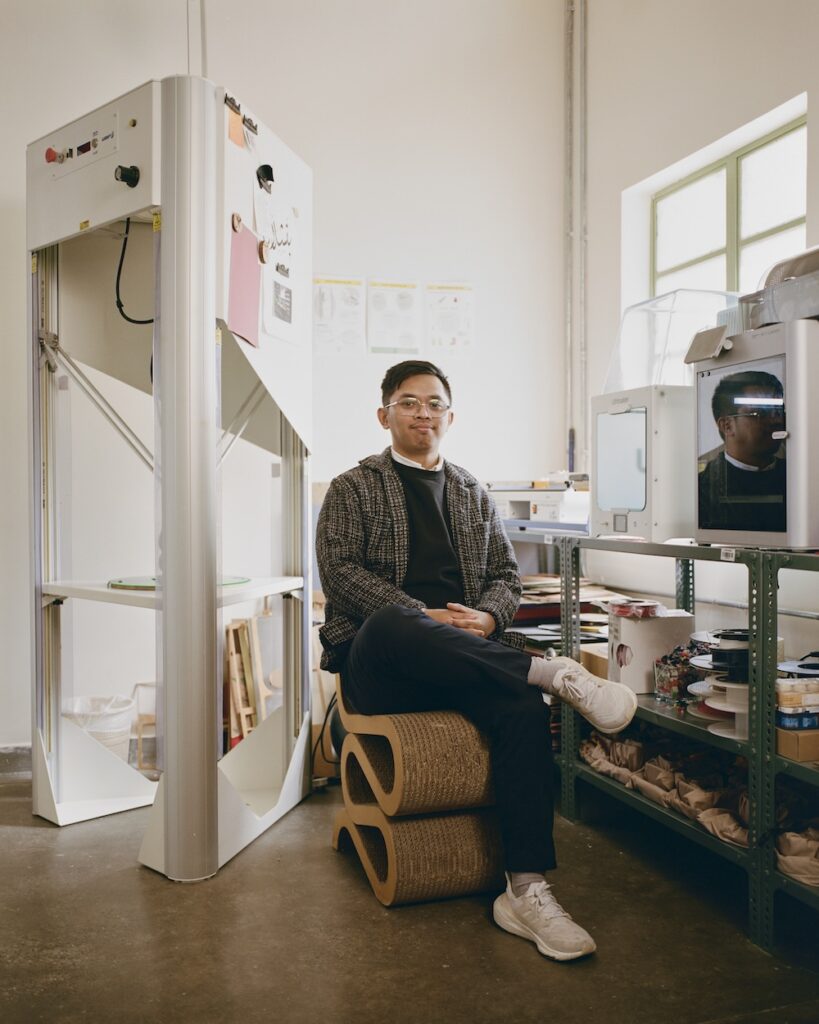 A man wearing glasses, a sweater, and sneakers sits on stacked cardboard rolls in a liwan-inspired workspace with 3D printers, shelves of supplies, and a window letting in daylight.