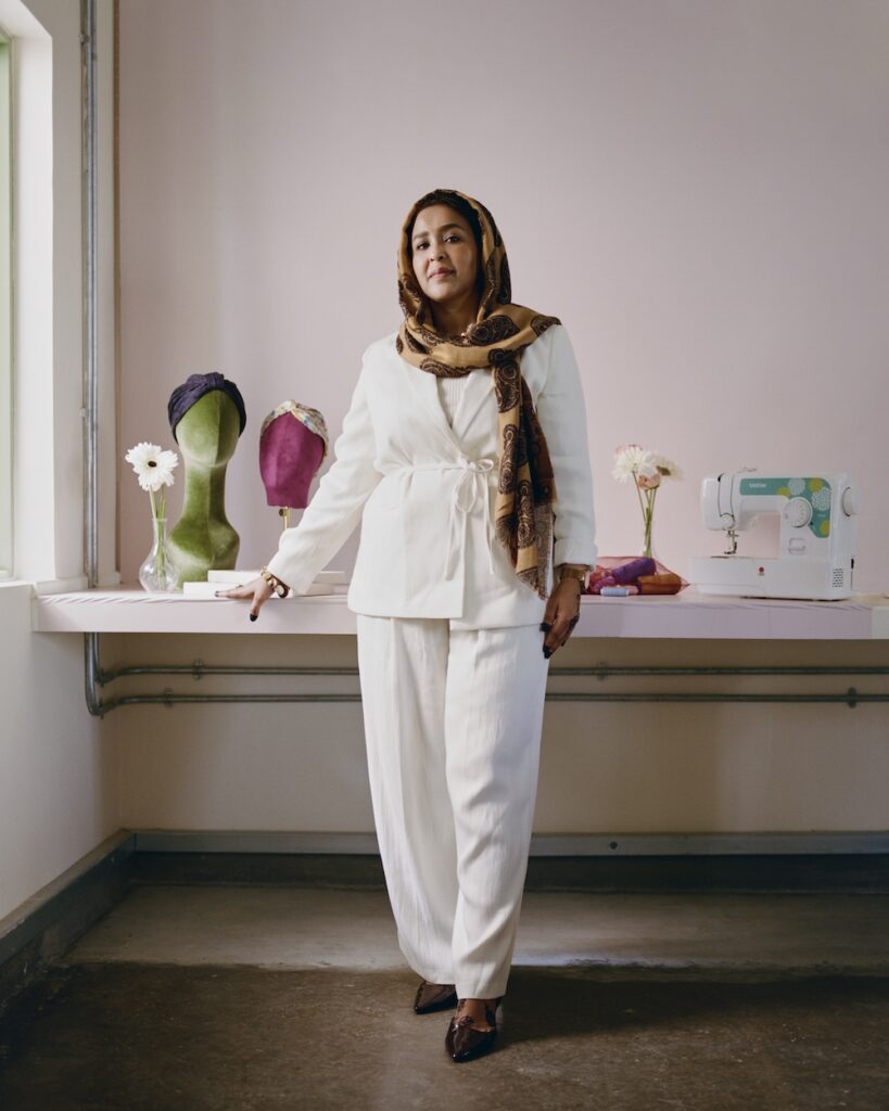 A woman wearing a white suit and patterned headscarf stands in a liwan by a table with mannequins in head coverings, two vases with flowers, folded fabric, and a sewing machine, against a pale pink wall.