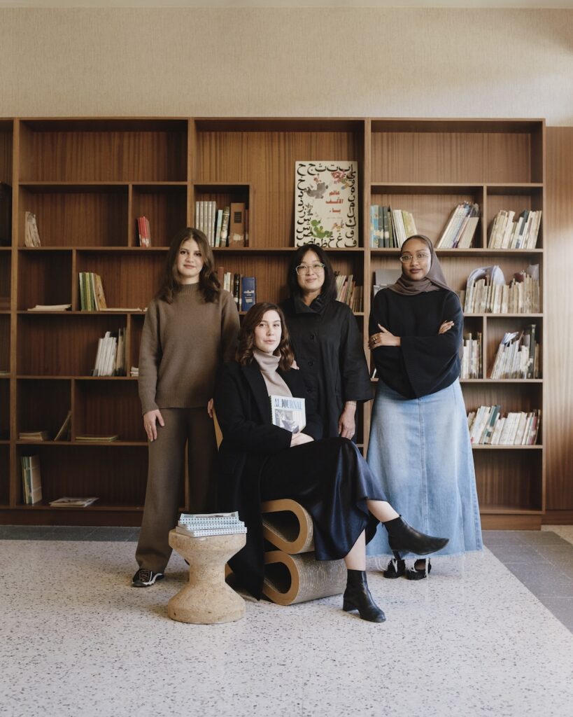 Four women pose in front of wooden bookshelves in a liwan. Three stand, while one sits holding a book. All are dressed in modest, neutral clothing, with shelves of books visible behind them.