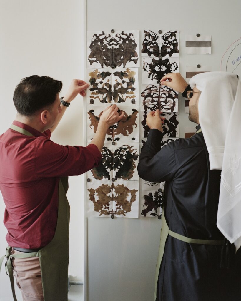 Two people in aprons pin inkblot images, resembling Rorschach tests, onto a whiteboard in a liwan-inspired setting. One wears a red shirt, the other a black robe with a white headscarf.