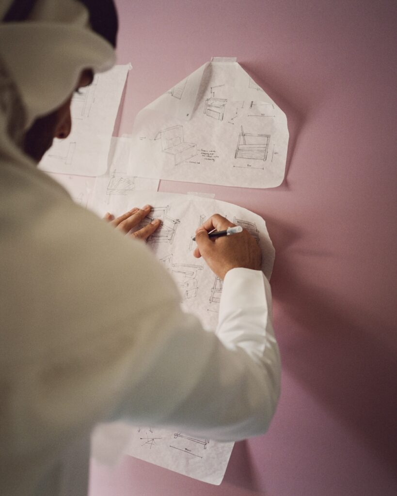 A person in traditional Middle Eastern clothing sketches on paper taped to a pink wall in a liwan, with several hand-drawn technical diagrams and notes visible.