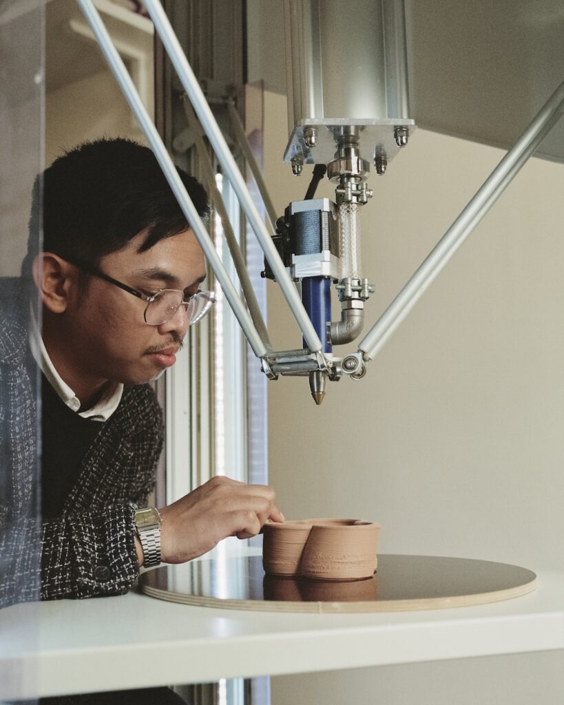 A person wearing glasses examines a clay object being shaped on a 3D printer at Liwan, focusing intently on the printing process.