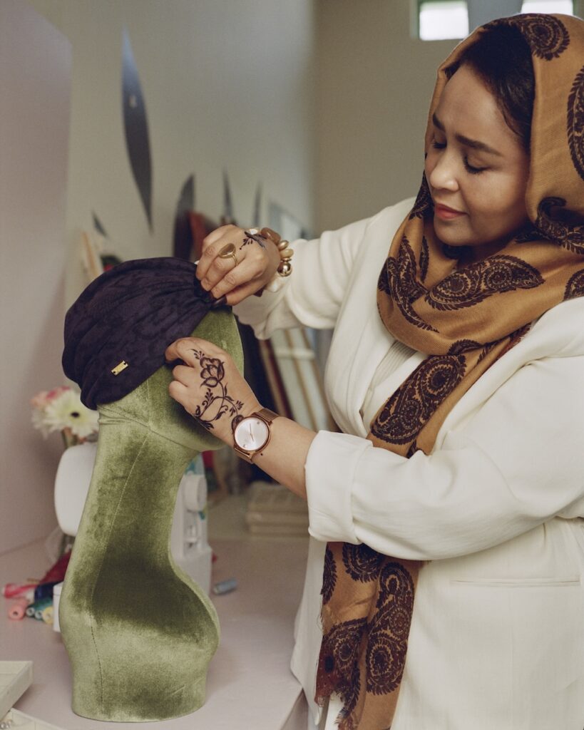 A woman wearing a patterned headscarf and white coat adjusts a dark-colored cap on a green mannequin head inside a liwan. She has intricate henna designs on her hand, a watch on her wrist, and she appears focused on her work.