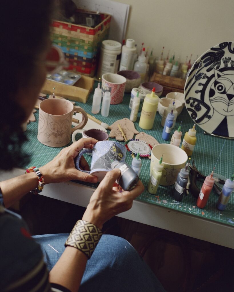 A person decorates a ceramic object with paint in the liwan, surrounded by colorful paint bottles, mugs, and a plate with a cat design on a worktable in an art studio.
