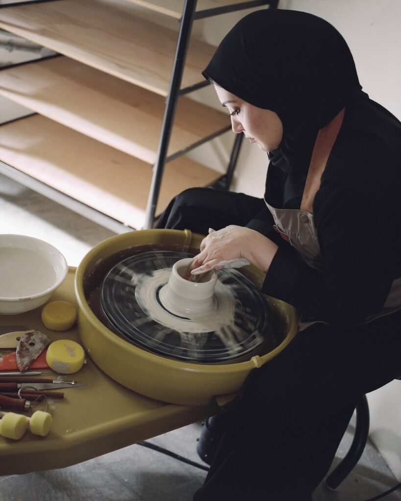A woman wearing a black hijab shapes clay on a pottery wheel. She is seated at a yellow table with pottery tools and sponges, crafting her piece in a liwan-inspired studio. Wooden shelves are in the background.