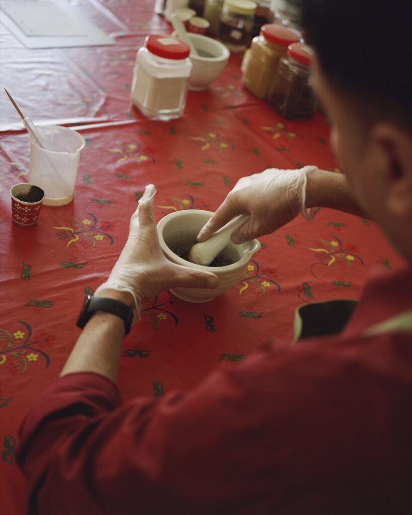 A person wearing gloves grinds ingredients in a mortar and pestle on a red patterned tablecloth, surrounded by jars and containers, evoking the warm ambiance of a liwan.