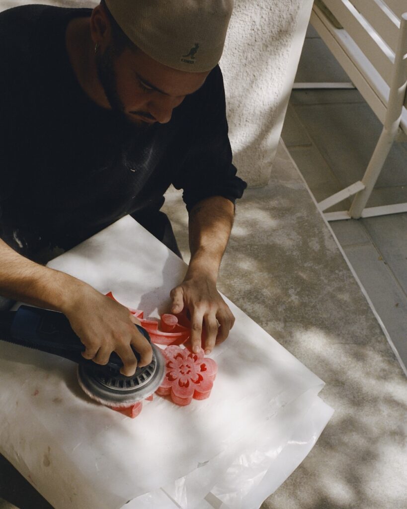 A person wearing a beige cap uses a power tool to sculpt or polish a liwan-inspired, flower-shaped object made of pink material on a white surface outdoors.