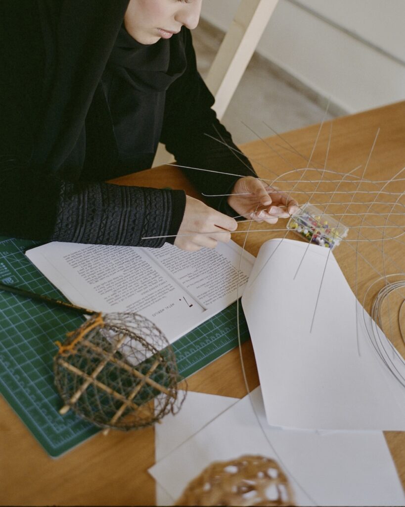 A person in a black hijab works on a wire sculpture at a desk in the liwan, surrounded by an open book, papers, a cutting mat, and two spherical wire models.