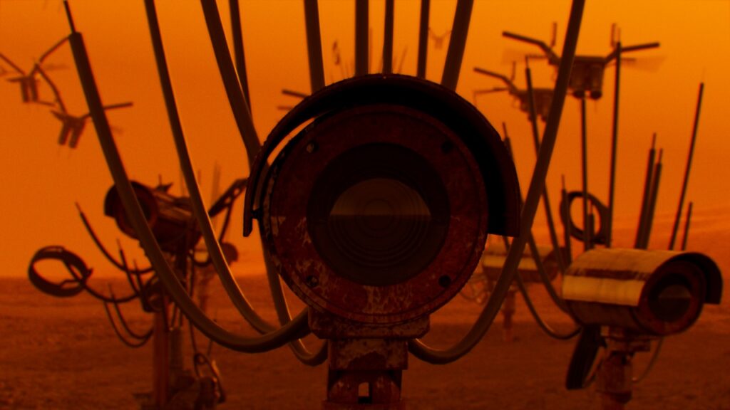 A close-up of a weathered mechanical device with a large circular lens, surrounded by curved metal rods, set against an orange, hazy sky. Similar structures in the background evoke a surreal, industrial scene reminiscent of gulf futurism.