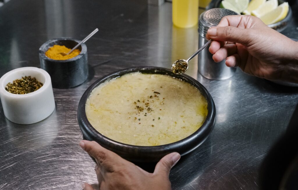 A person holds a black liwan bowl of soup or porridge on a metal counter, sprinkling a spice on top with a small spoon; bowls of spices and lime wedges are nearby.